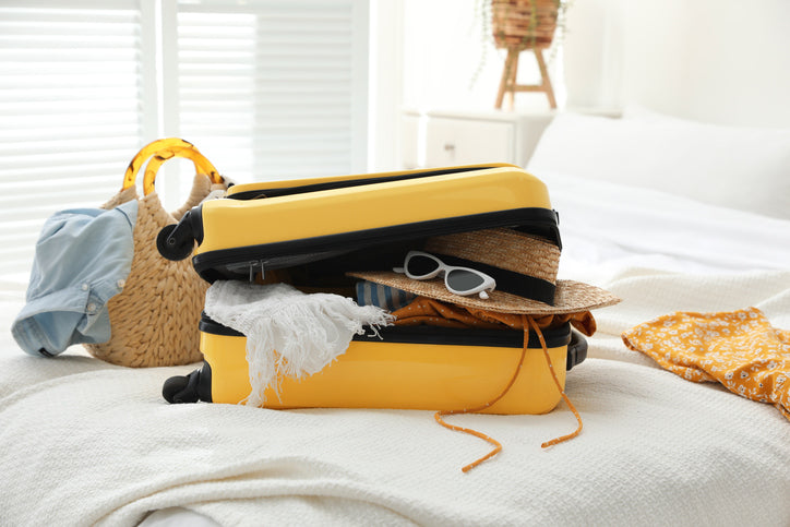 A half-open yellow suitcase full of clothes placed on a bed together with a beach bag