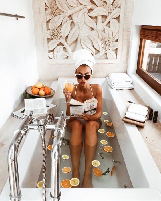 A girl bathing in a white tub, holding a book in her left hand and an orange juice in her right hand. A white towel wraps her hair and she wears sunglasses.