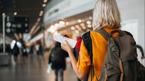 A blonde-haired girl, from behind, with a yellow sweater and a brown backpack, holding a plane ticket in her left hand while at the airport