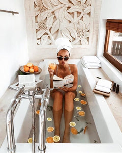 A girl bathing in a white tub, holding a book in her left hand and an orange juice in her right hand. A white towel wraps her hair and she wears sunglasses.